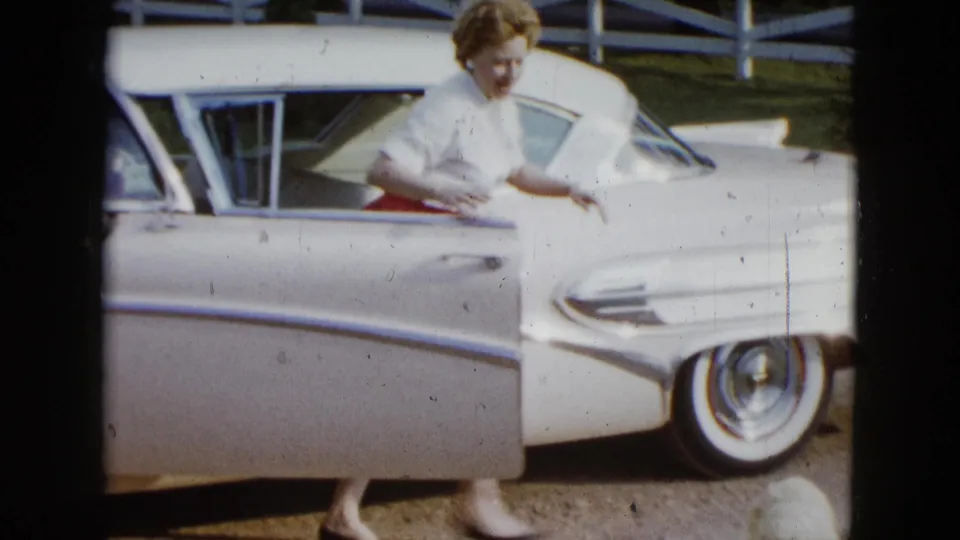 1958 Las Vegas frame showing a woman exiting a white vintage sedan while a small white dog approaches near the lower edge.