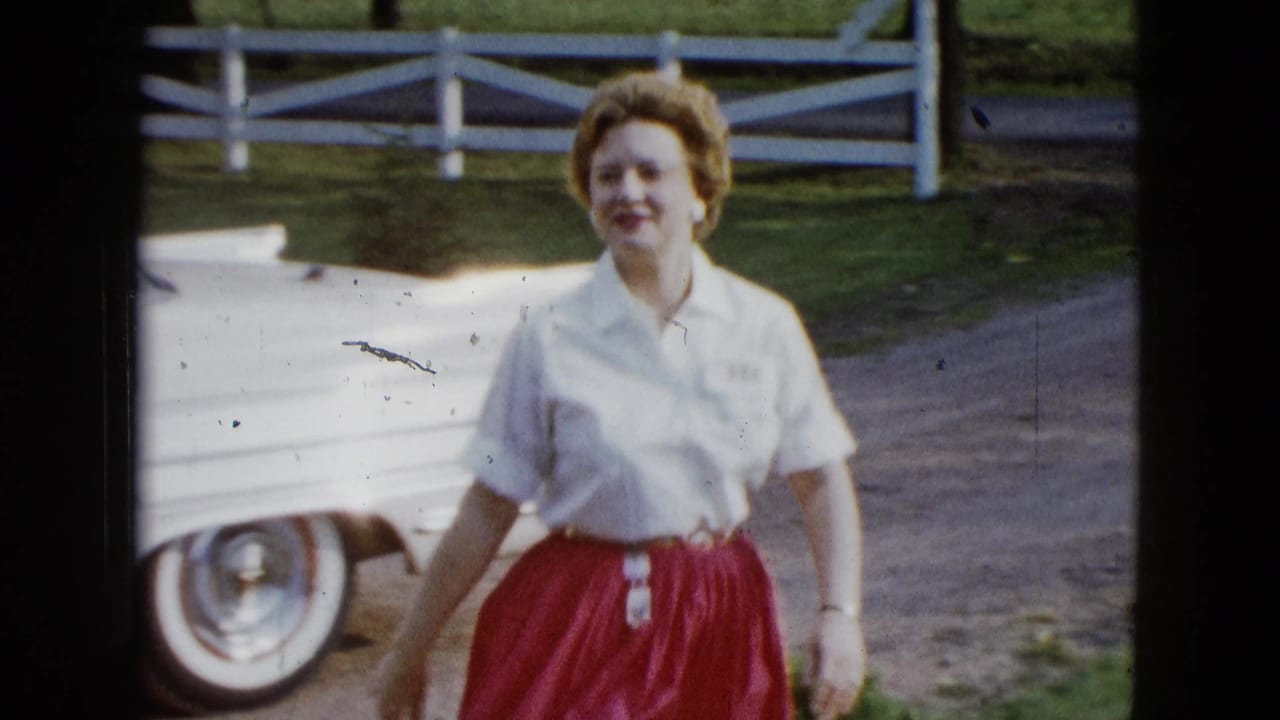 The woman smiles while walking toward the camera, with a white 1950s car and fence behind her in Las Vegas footage.