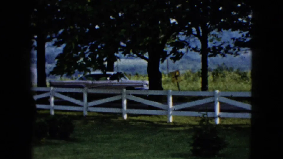 A white fence and shaded lawn frame a distant 1950s car, with trees and hills or fields beyond.