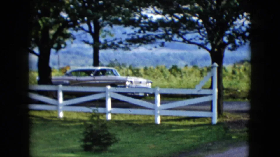 A 1950s automobile appears beyond a white fence under trees, with open landscape visible behind it.