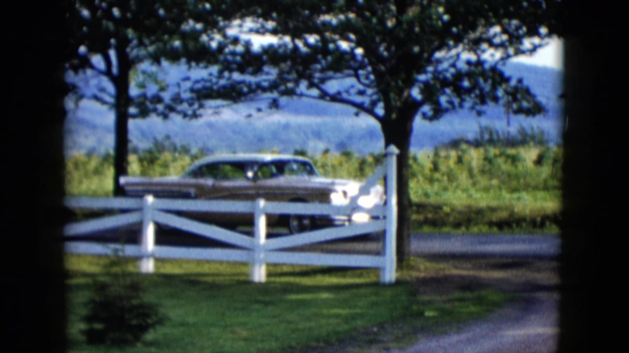 Two-tone vintage sedan passes a white fence beside trees and distant hills.