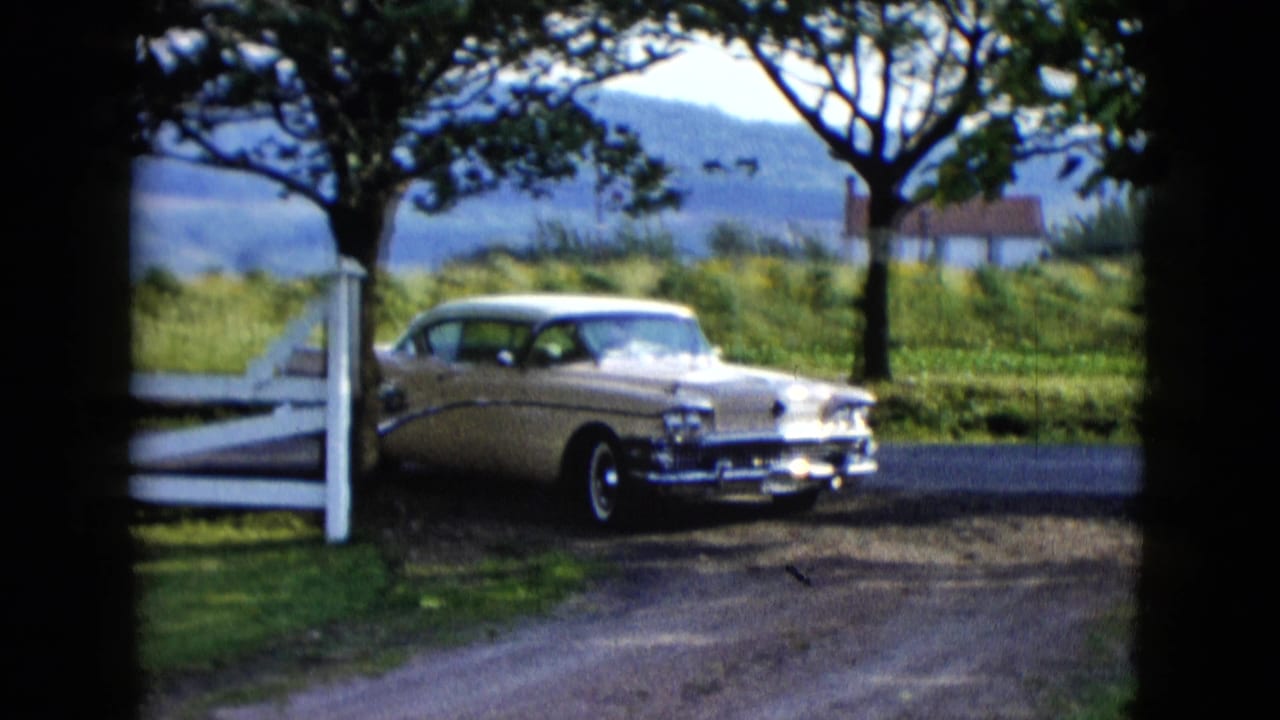 The classic sedan turns from the paved road near a white fence, with grass, trees, hills, and a small distant house behind it.