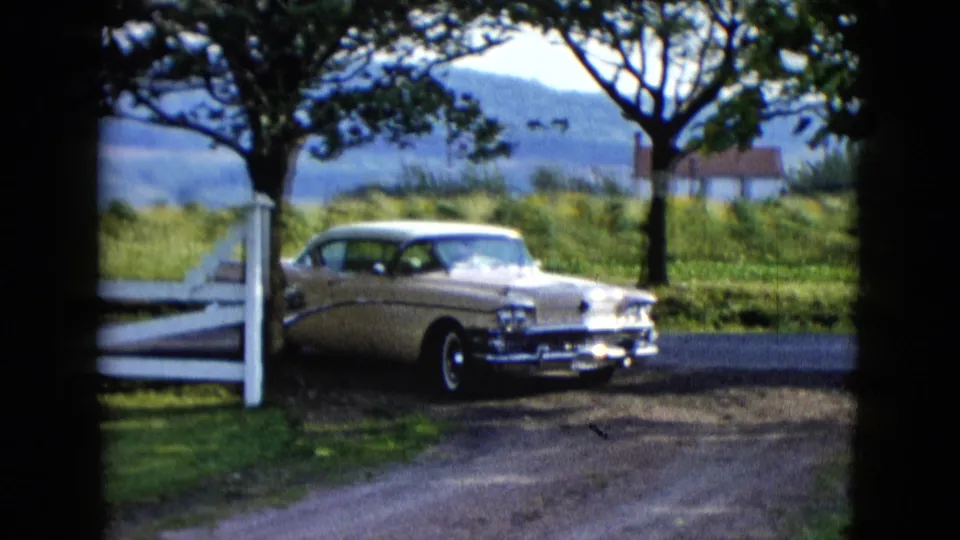The classic sedan turns from the paved road near a white fence, with grass, trees, hills, and a small distant house behind it.