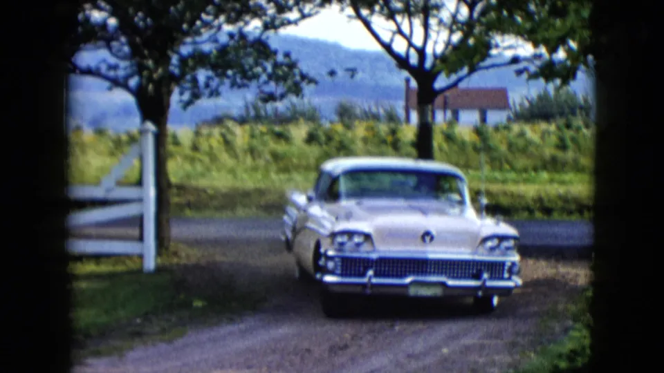 Front view of the two-tone sedan as it turns onto the dirt road, framed by a white fence, trees, grass, and blue hills.