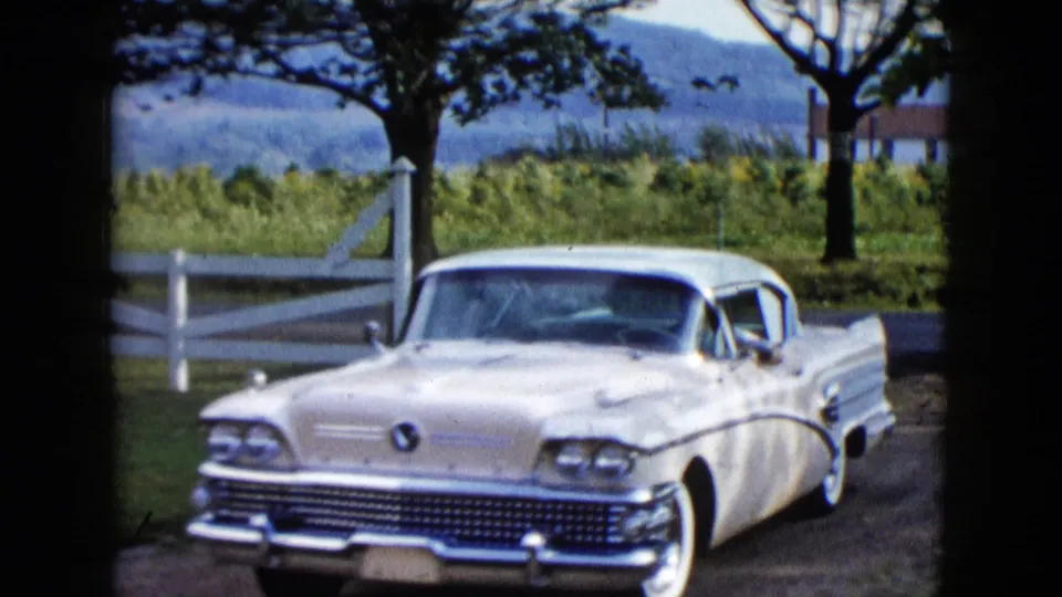 Close 1958 frame of the two-tone sedan on the dirt road, showing chrome trim, tailfin styling, white fence rails, and roadside greenery.