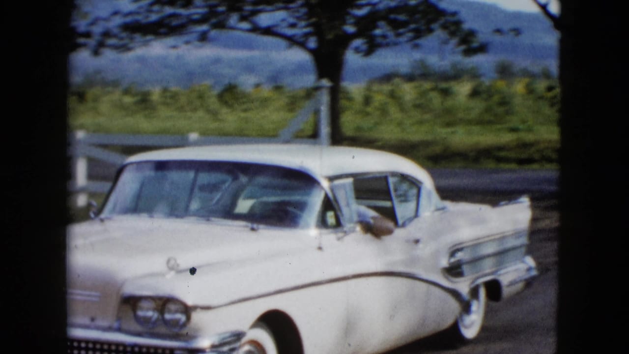 White 1950s car turning near a white fence and trees.
