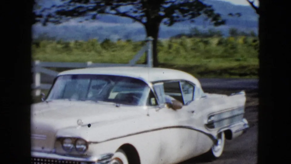 White mid-century American car in a rural Las Vegas-area driveway scene, with a fence, trees, and distant hills visible.