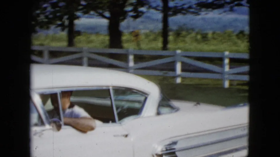 Adult man in a light shirt drives a white 1950s car past a fence and trees in the Las Vegas-area home-movie footage.