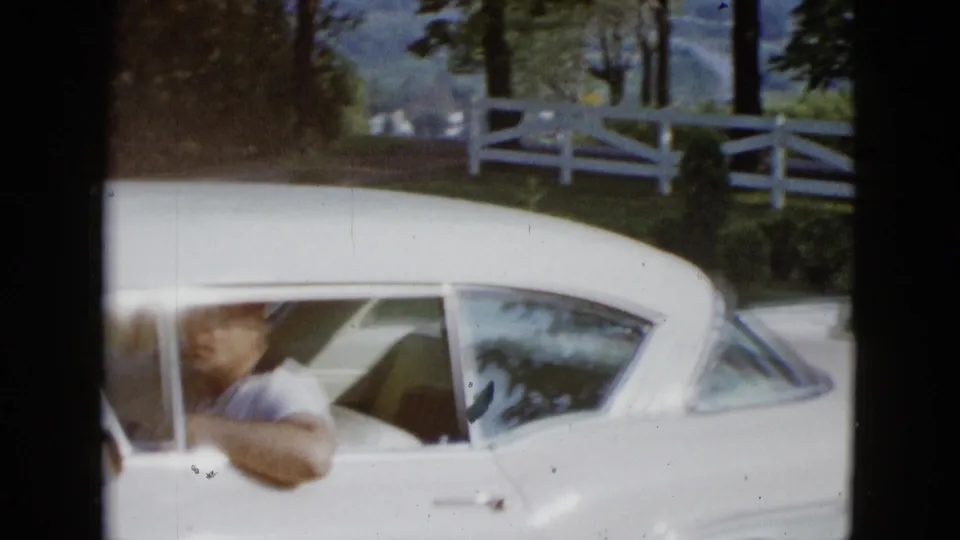 Close home-movie view of the adult driver inside a white classic car, with the rural fence and trees behind him.