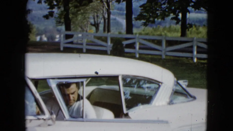 Adult man looks out from the driver seat of a white 1950s car near a white fence, trees, and distant hills.