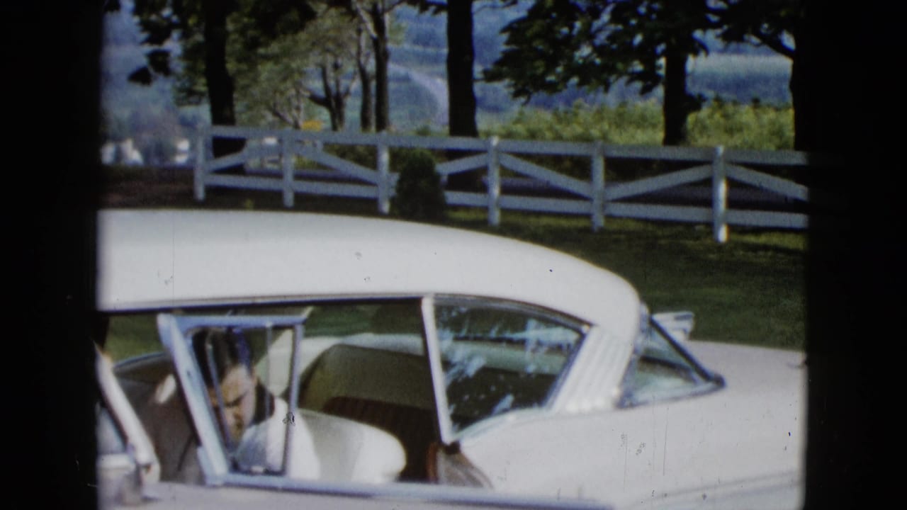 Man partly visible through the open door of a white classic car near a white fence and trees.
