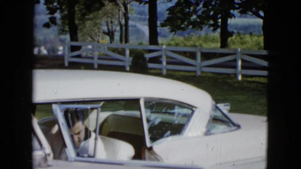 In 1958 Las Vegas-area home-movie footage, a man leans inside a white classic car with a white fence, trees, and distant hills behind him.