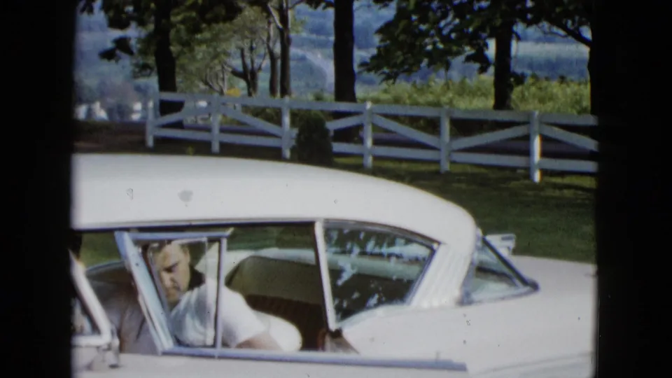 The man remains by the open door of the white 1950s car, with a white fence and shaded trees visible in the Las Vegas-area background.