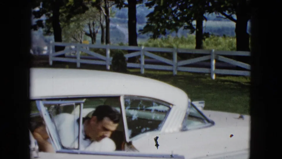 A man in a white shirt bends near the open door of the white car, framed by a fence, trees, and bright outdoor light.