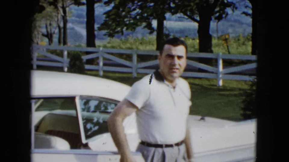 In the clearest frame, the man stands beside the white car wearing a white polo and light trousers, with fence, trees, and hills behind him.