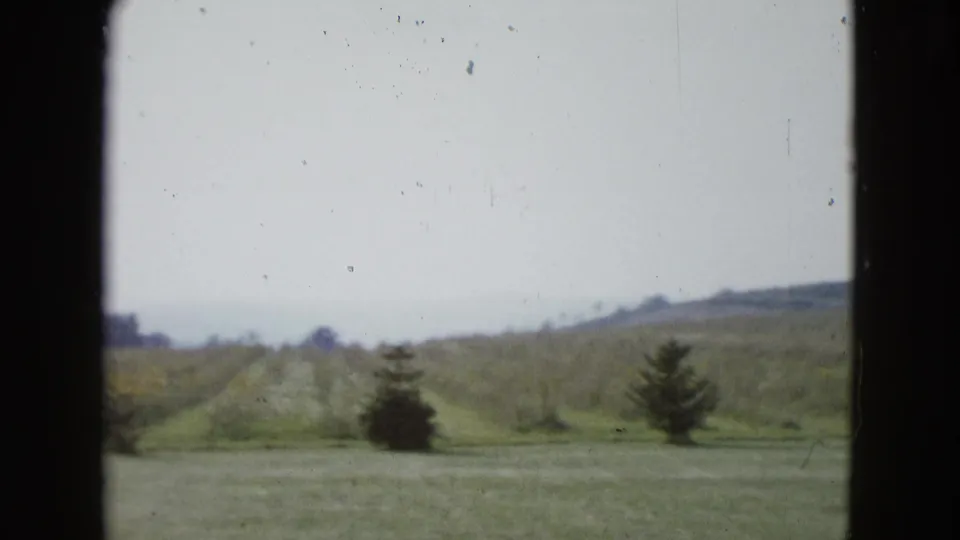1958 8mm frame from Las Vegas showing open grass, small trees, distant low hills, and a pale sky.
