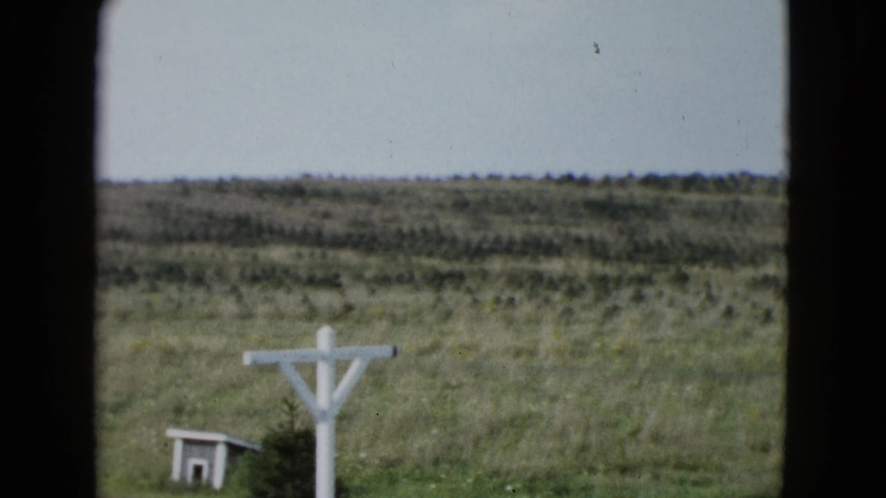1958 Las Vegas-area 8mm frame showing a rural field, distant scrub hills, a white wooden cross, and a small shed viewed through dark bars.