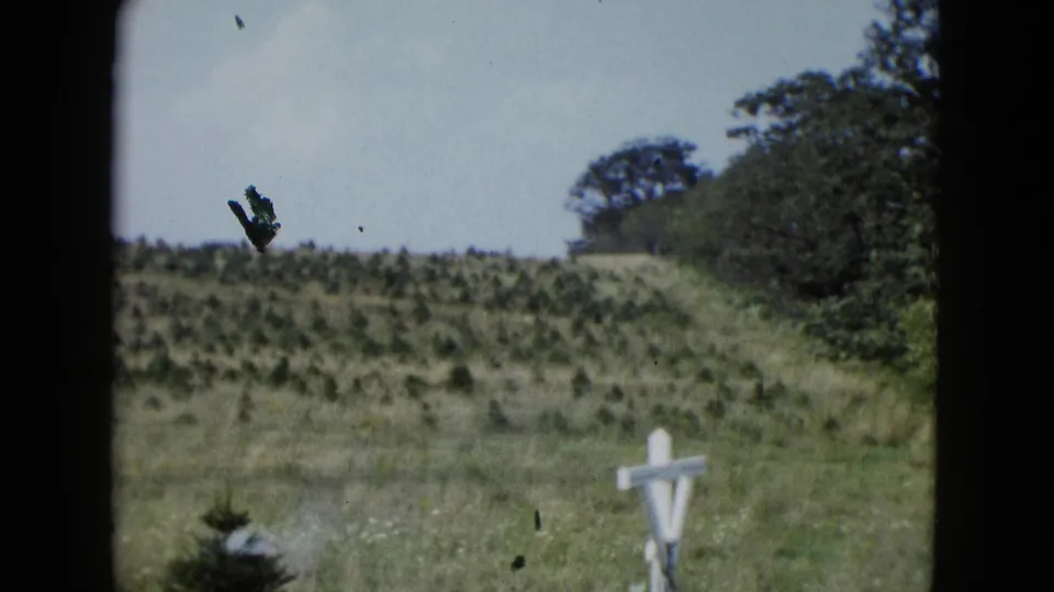 1958 Las Vegas-area 8mm frame showing a white wooden cross, open field, scrub hill, and trees along the right edge.