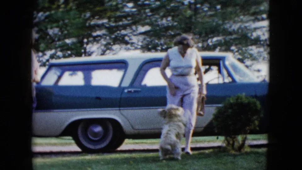 A woman in a light dress or outfit approaches a white dog beside a parked station wagon in 1958 Las Vegas footage.