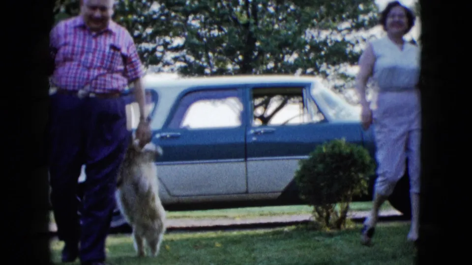 1958 Las Vegas home-movie frame showing a man beside a small dog while a woman walks past the parked station wagon.