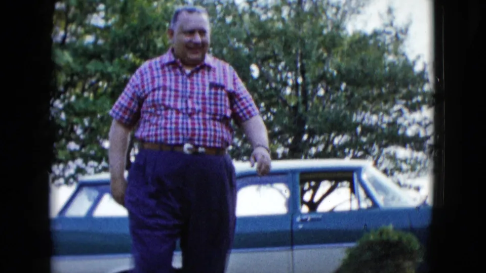 1958 Las Vegas frame of a smiling man in a red checkered shirt walking on a lawn with a blue and white station wagon behind him.
