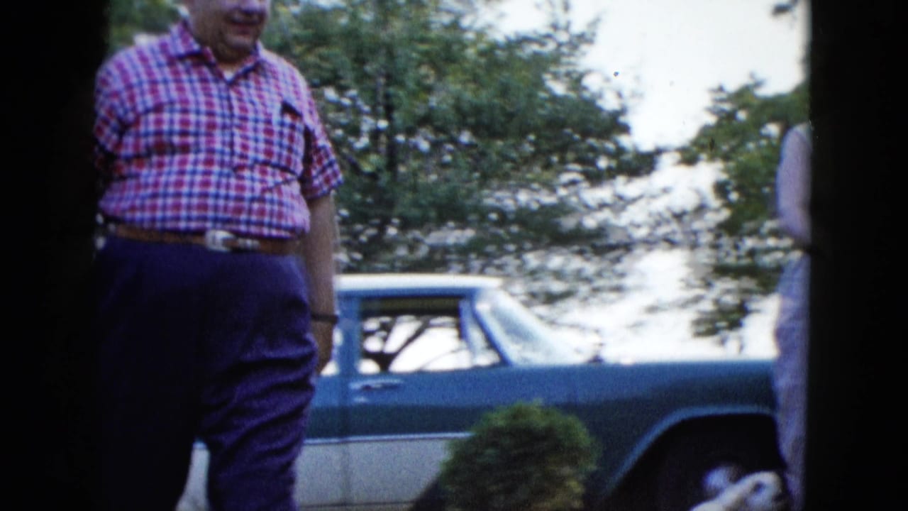 In a 1958 Las Vegas home-movie frame, a man in a red checkered shirt approaches near a two-tone car and leafy yard.