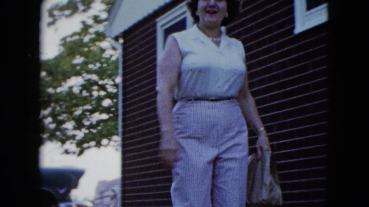 A woman in light sleeveless clothing walks near a brick house exterior in the stated 1958 Las Vegas home-movie setting.