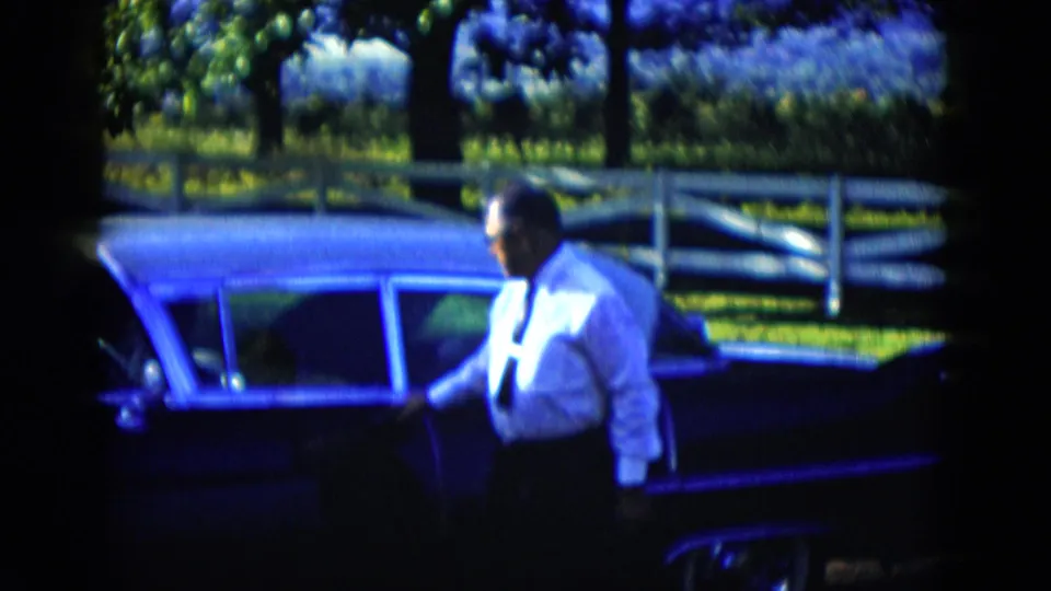 A man in a white shirt and tie holds the door of a dark car in a rural-looking outdoor scene from the home-movie clip.