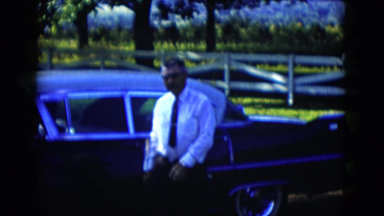 In 1958 Las Vegas-area 8mm footage, a man in glasses stands near a dark midcentury sedan with grass, trees, and a white fence behind him.
