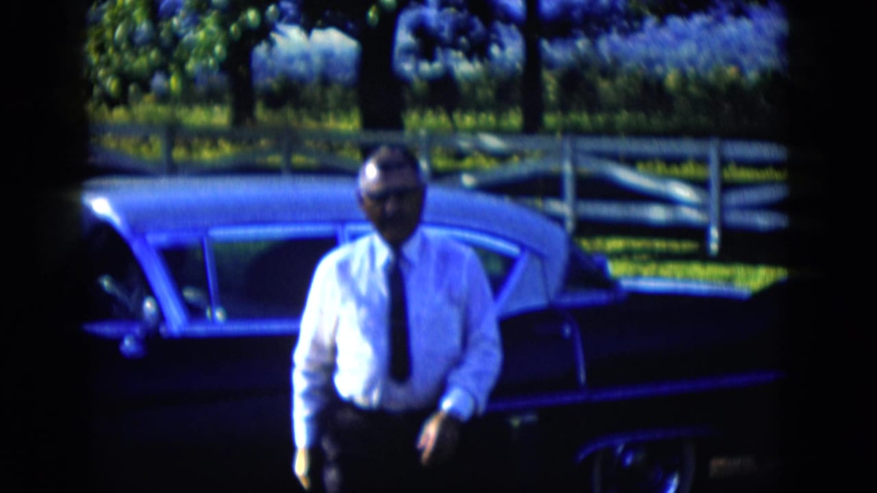 The man moves away from the parked sedan in 1958 Las Vegas-area home-movie footage, with a white fence and leafy background visible.
