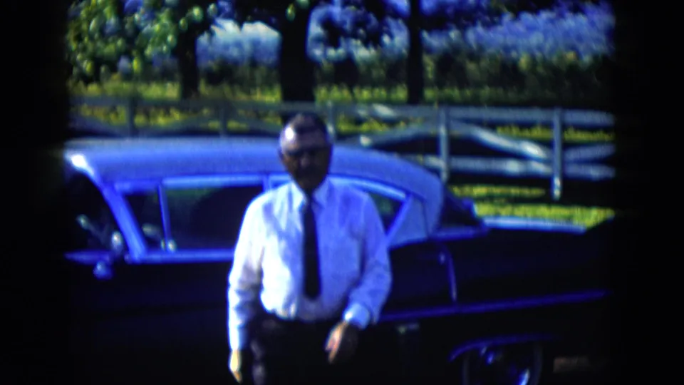 The man moves away from the parked sedan in 1958 Las Vegas-area home-movie footage, with a white fence and leafy background visible.