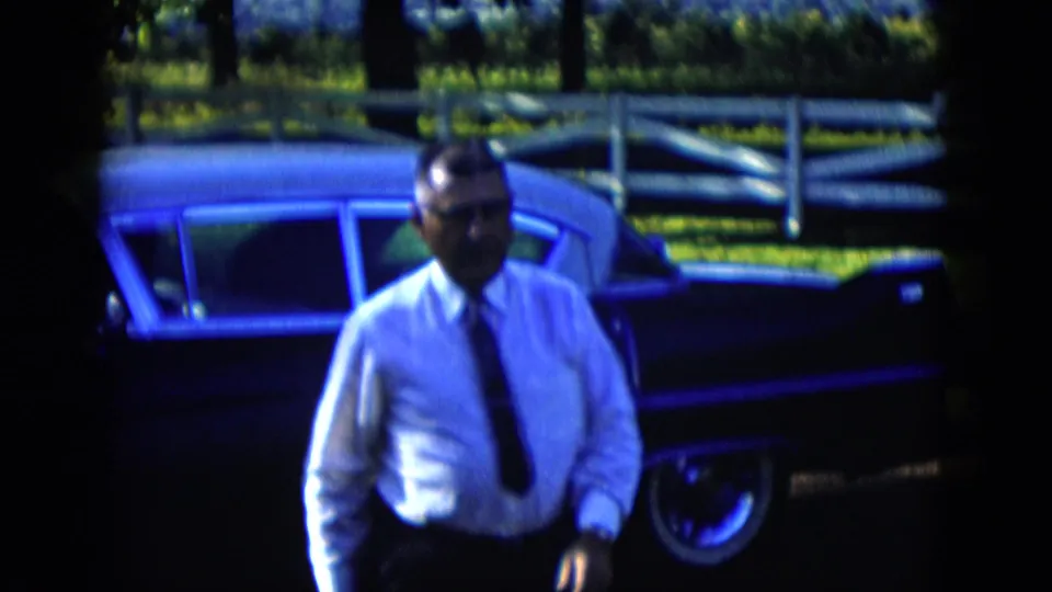A closer frame shows the bespectacled man walking past the dark sedan in 1958 Las Vegas-area 8mm footage, with fence and greenery behind.
