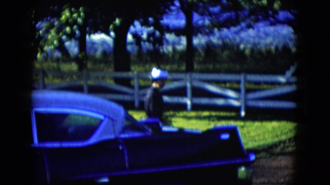 A dark vintage car sits by a white rail fence as a woman in a dark dress and light hat stands nearby in the Las Vegas-area setting.