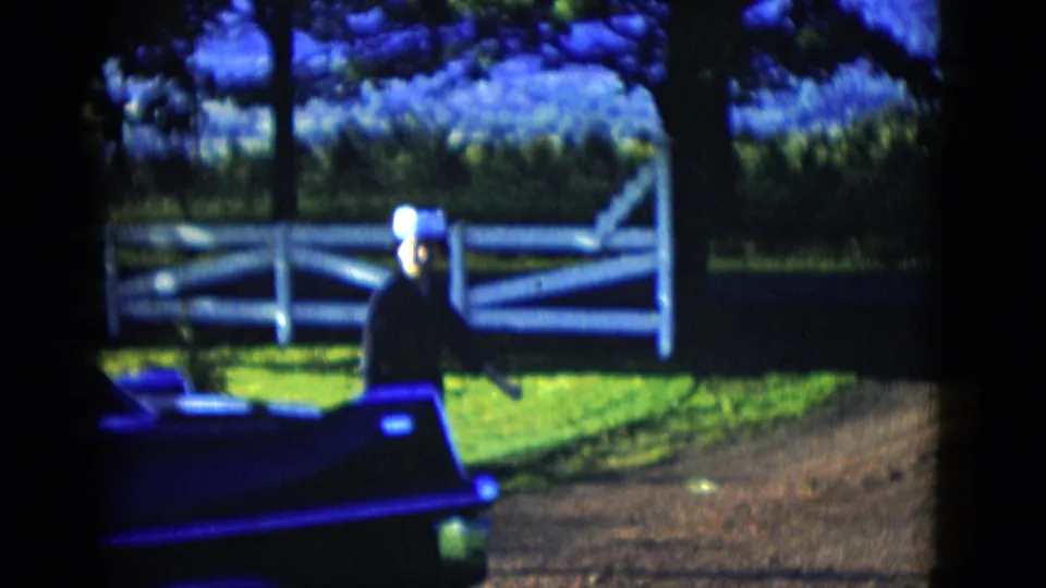 The woman waits beside the dark car near a white rail fence, with trees and bright grass behind her in the 1958 home movie.