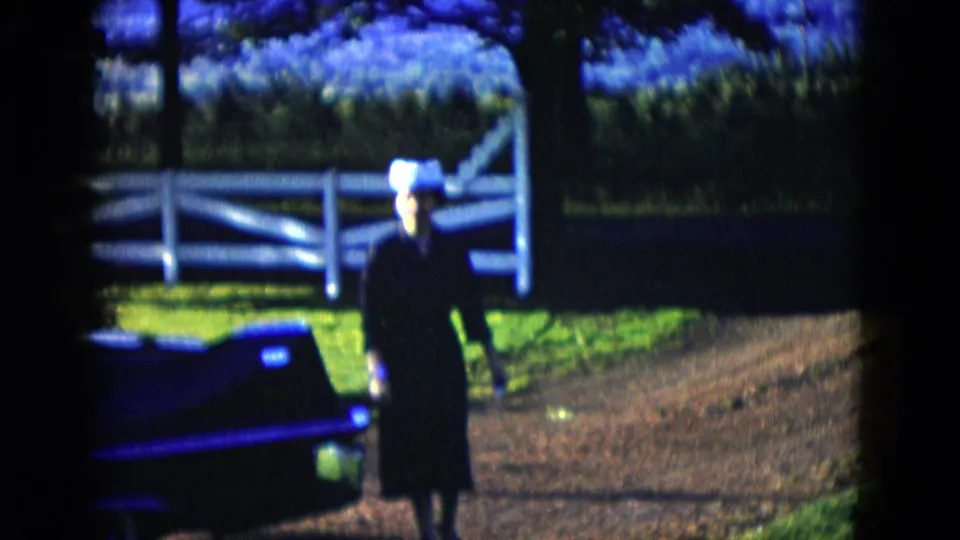 The woman walks on the dirt ground near the dark vintage car and white fence, continuing the quiet meeting moment.