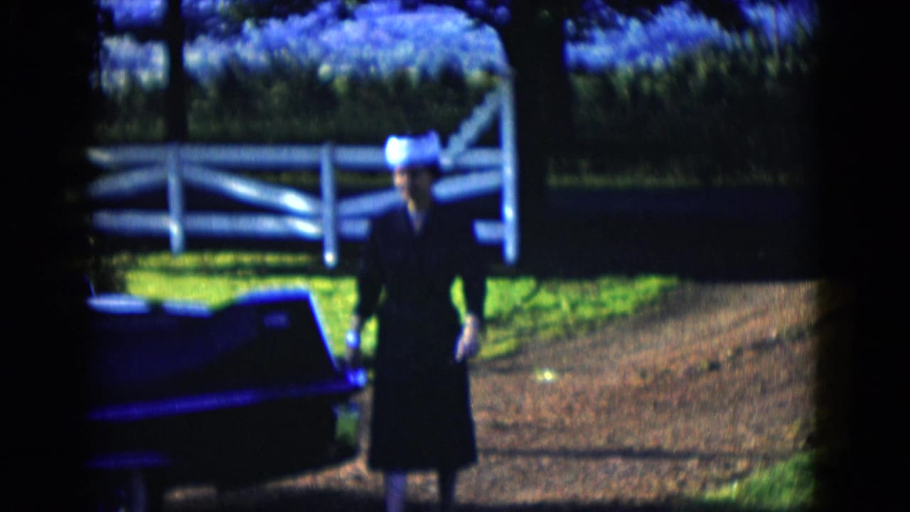In 1958 Las Vegas home-movie footage, a woman in dark clothing and a light hat walks beside a dark classic car near a white fence.