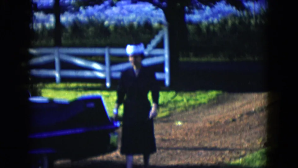 In 1958 Las Vegas home-movie footage, a woman in dark clothing and a light hat walks beside a dark classic car near a white fence.