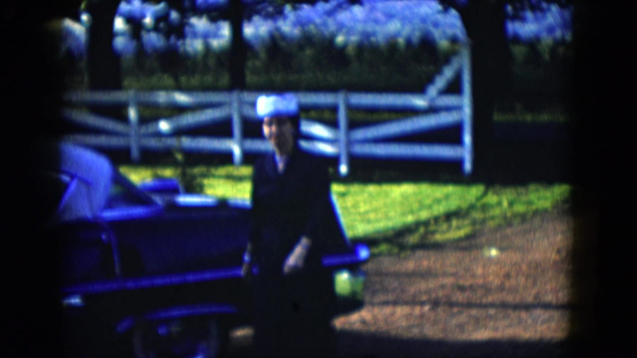 The woman moves closer to the camera beside the vintage car, with white fence rails, lawn, and shrubs visible in bright sunlight.