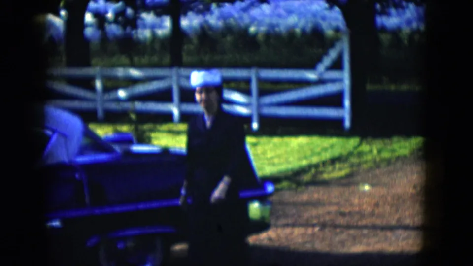 The woman moves closer to the camera beside the vintage car, with white fence rails, lawn, and shrubs visible in bright sunlight.