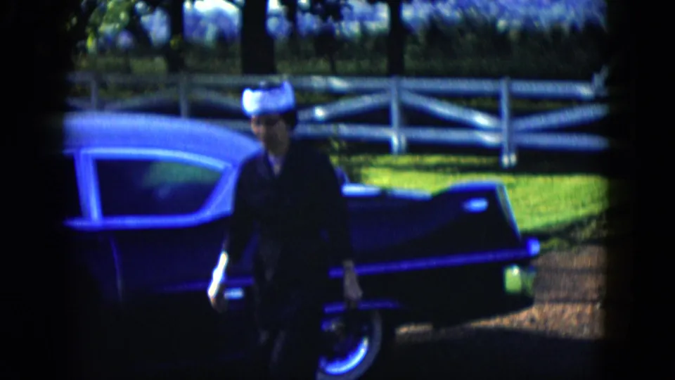 A closer view shows the woman passing the side of the dark classic car, framed by the sunlit lawn and white garden fence.