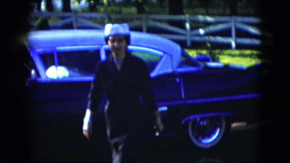 The woman is nearest the camera at the end of the clip, with the dark classic car, white fence, lawn, and shrubs behind her.