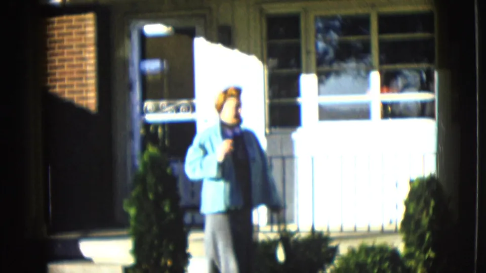 1958 Las Vegas frame of a woman in a blue jacket standing near a residential entrance with windows, shrubs, and strong daylight.