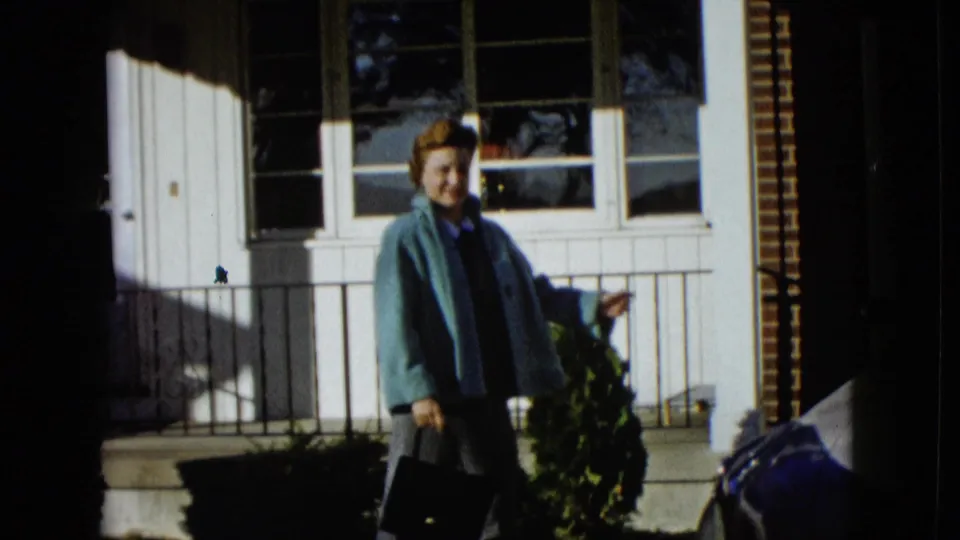 Woman in a blue jacket stands outside a Las Vegas residence in a 1958 home-movie frame, with windows and shrubs behind her.