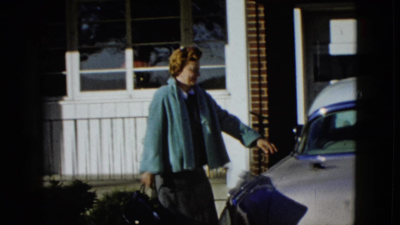 Woman in a teal jacket walks toward a pale classic car outside a sunny house.