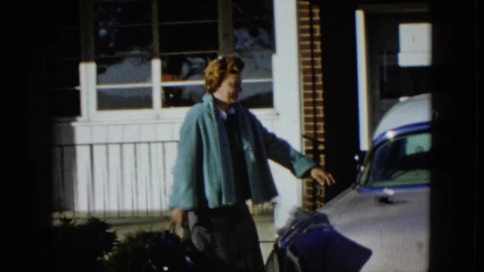 In supported 1958 Las Vegas home-movie footage, a woman carries a handbag toward a pale classic car in front of a suburban house.