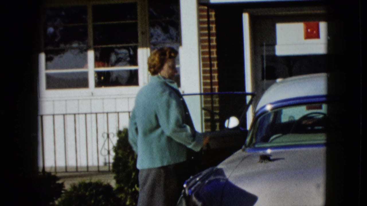 Woman stands beside the driver-side door of a pale classic car near a house.
