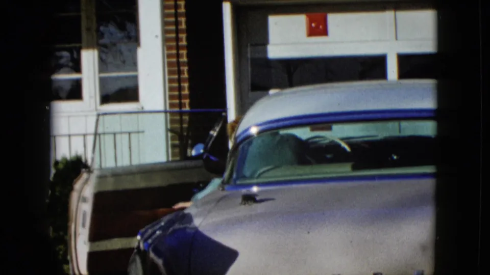 The woman leans into the open driver-side doorway of the pale classic car, partly obscured by the vehicle in bright sun.