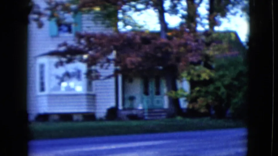 1958 Las Vegas frame of a white residential house with green shutters, front lawn, trees, and road in the foreground.