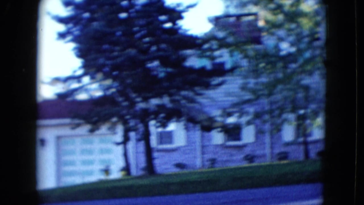 1958 Las Vegas residential view with a pale garage door, adjoining house, evergreen tree, sloped lawn, and street edge.