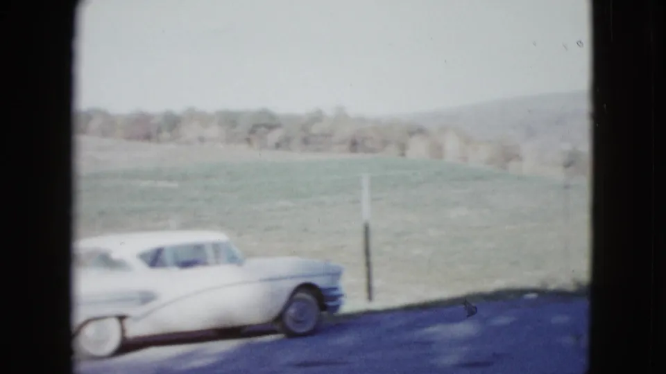 1958 8mm frame near Las Vegas showing a white 1950s sedan at the roadside with open fields and low hills behind it.